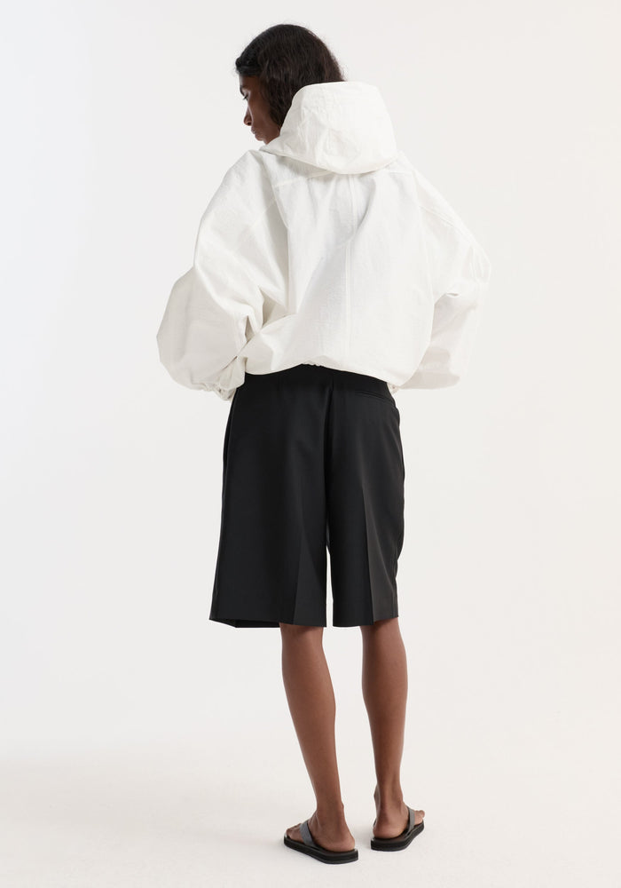 Person in optic white textured cotton-blend anorak with hood, front and side pockets, adjustable straps, paired with tailored black shorts and slide sandals.