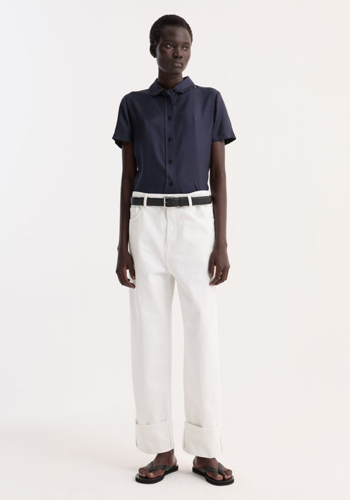 Person wearing Embroidered silk fitted shirt with short sleeves and embroidery, tucked into high-waisted trousers, accessorized with a black belt and sandals.