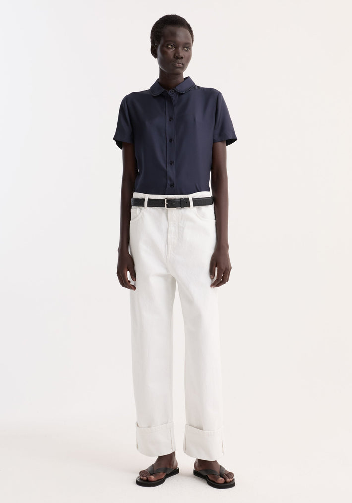 Person wearing Embroidered silk fitted shirt with short sleeves and embroidery, tucked into high-waisted trousers, accessorized with a black belt and sandals.