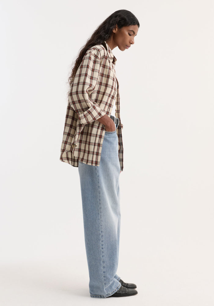 Young person in a beige-and-brown plaid overshirt, hands in pockets, wearing high-waisted jeans and black slip-on shoes. Model's long hair flows down their back.