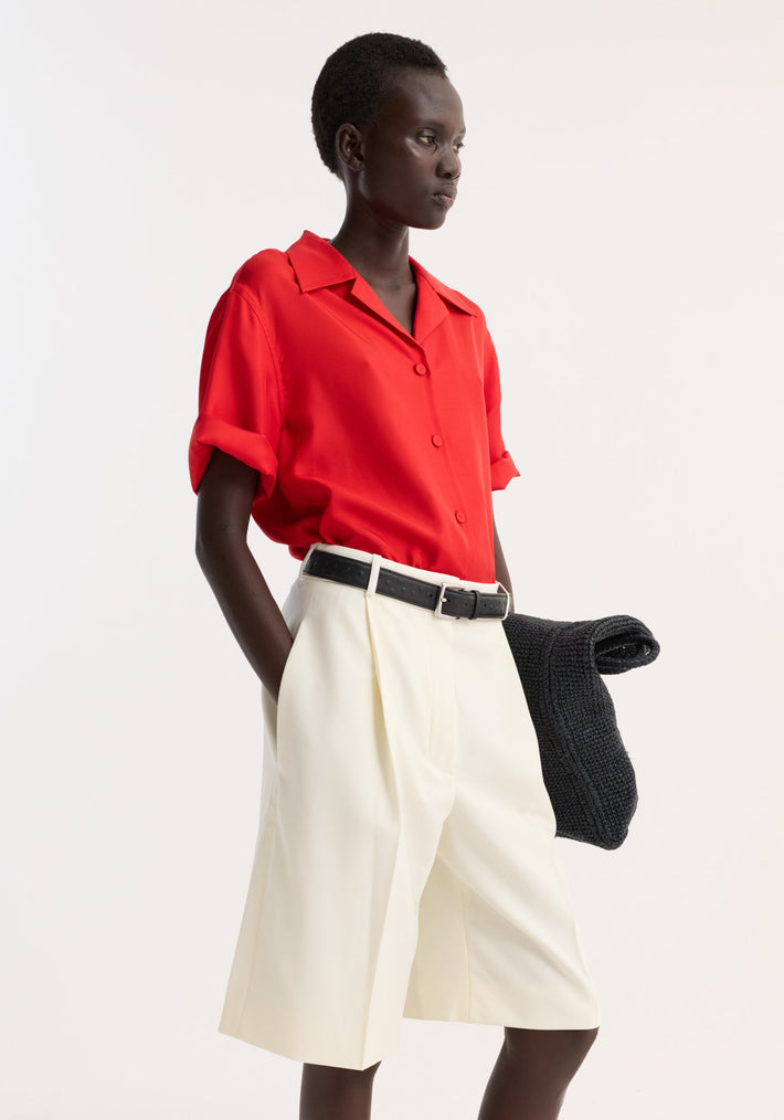 Dark-skinned model wears a Camp collar silk shirt in red, featuring short sleeves and dropped shoulders, paired with tailored shorts and a black belt.