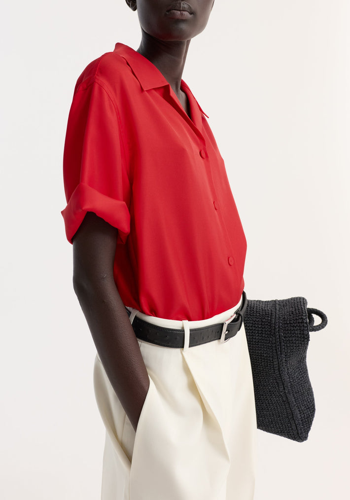 Person wearing a red Camp collar silk shirt with rolled sleeves, tucked into cream trousers, holding a black tote bag.