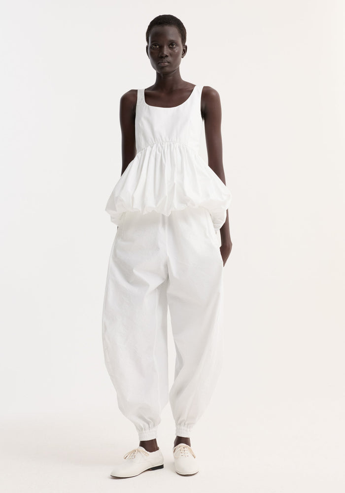 Model wears Voluminous peplum top | optic white, featuring a sleeveless scoop neck, low back, and sculptural peplum, paired with loose harem-style pants.