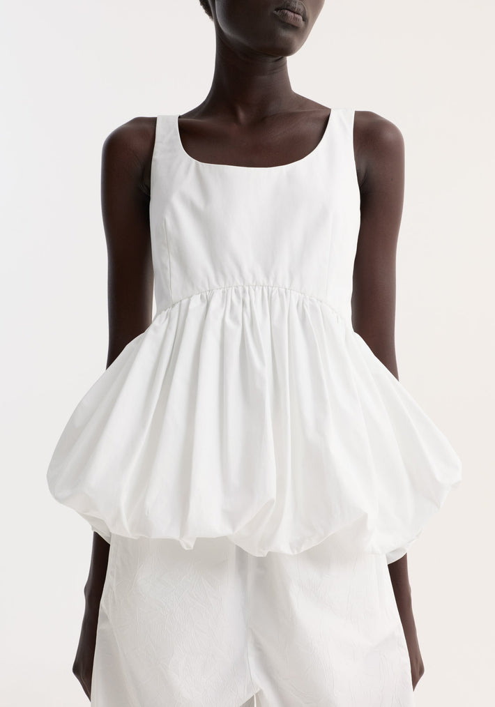 Voluminous peplum top in optic white, featuring a sleeveless scoop neck, low back, and sculptural peplum, worn by a dark-skinned model against a neutral background.
