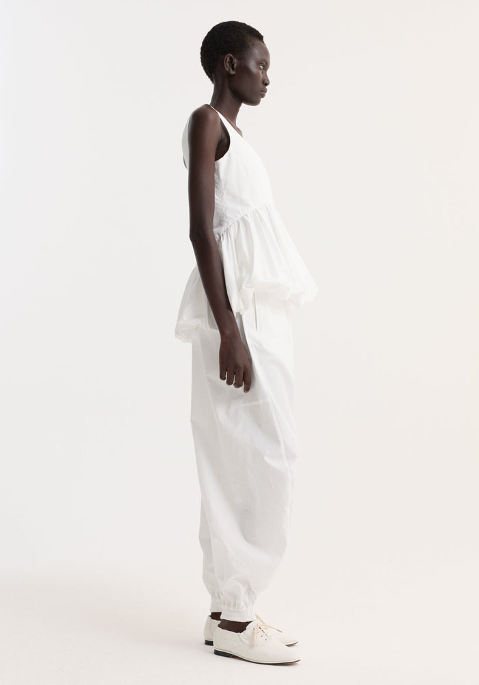 Voluminous peplum top | optic white on a slender person in profile, highlights a scoop neck, sleeveless design, and sculptural peplum detailing, enhancing the waist.