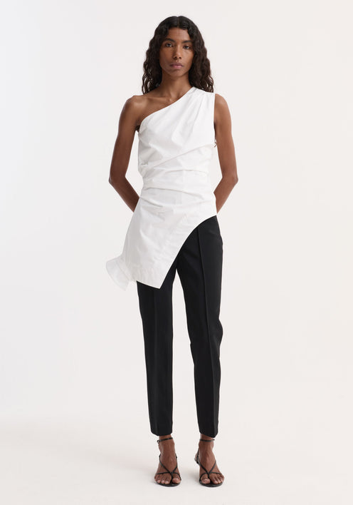 Model in Reimagined one shoulder top, optic white, with asymmetric drape and inverted collar, paired with tailored black trousers and strappy sandals, hands behind back.