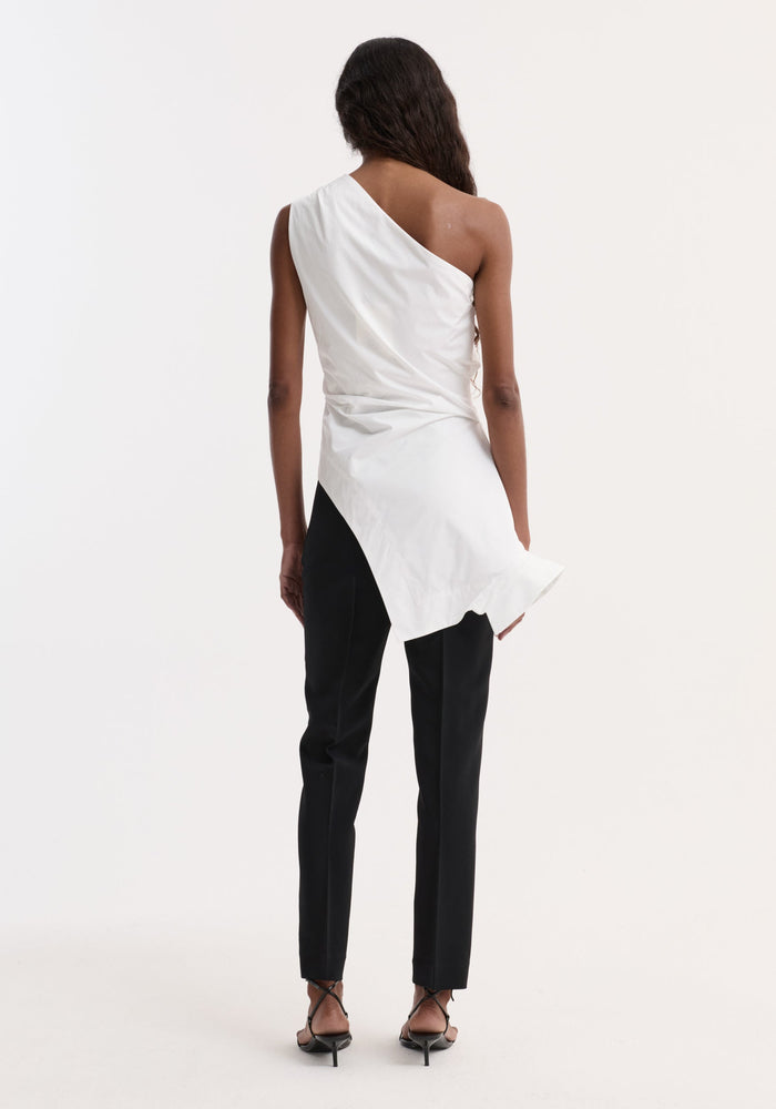 Back view of model wearing the Reimagined one shoulder top in optic white, featuring an asymmetric drape, fitted waist, and deconstructed detailing with slim-fit black trousers.