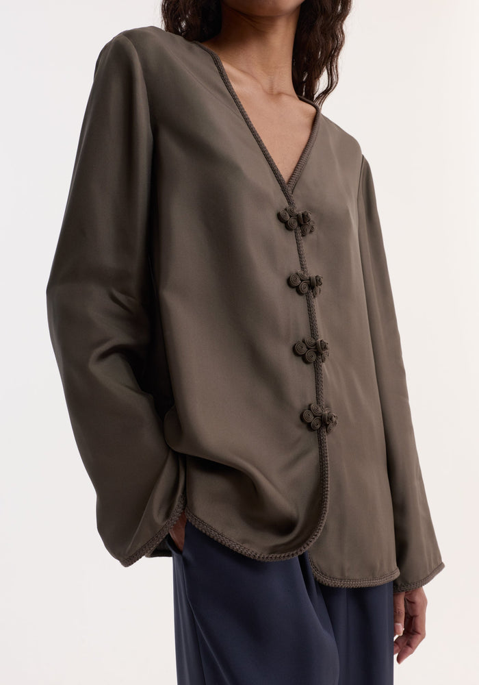 Person wearing a Pankou closure silk top | earth, featuring a V-neck, frog knot closures, embroidered edges, and long sleeves, paired with dark navy trousers.