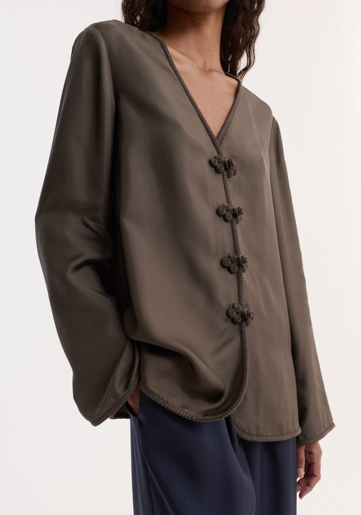 Person wearing a Pankou closure silk top | earth, featuring a V-neck, frog knot closures, embroidered edges, and long sleeves, paired with dark navy trousers.