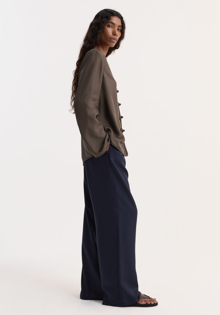 Pankou closure silk top with embroidered edges, shown in profile. The model wears it with navy wide-leg trousers and black flip-flops, highlighting its relaxed fit.