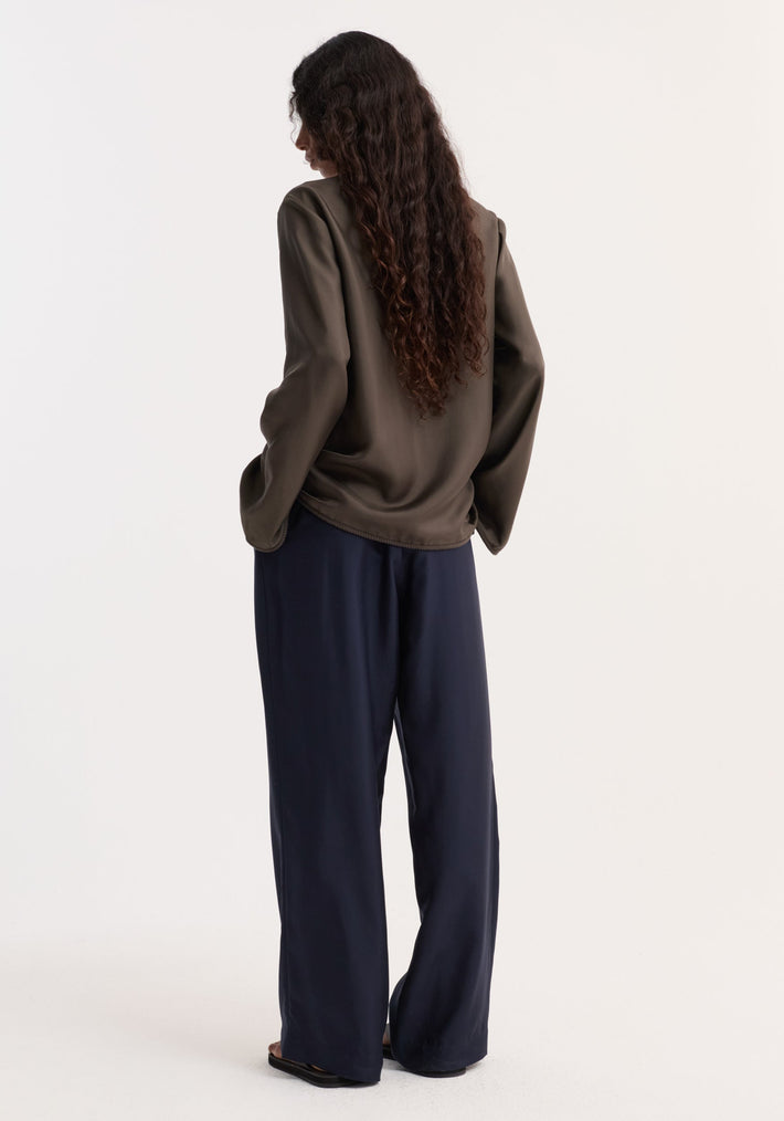 Person in olive Pankou closure silk top with embroidered edges, hands in navy trousers pockets, showcasing relaxed, minimalist style.