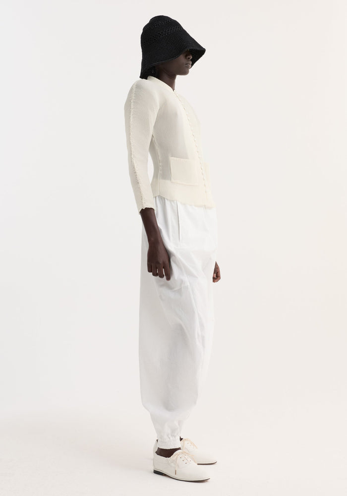 Model wearing Plissé cardigan | cream, featuring a ribbed texture, stand-up collar, fabric-covered buttons, and patch pockets, styled with white trousers and a black crochet hat.