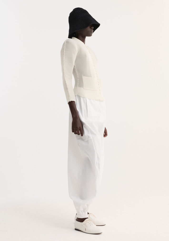 Model wearing Plissé cardigan | cream, featuring a ribbed texture, stand-up collar, fabric-covered buttons, and patch pockets, styled with white trousers and a black crochet hat.