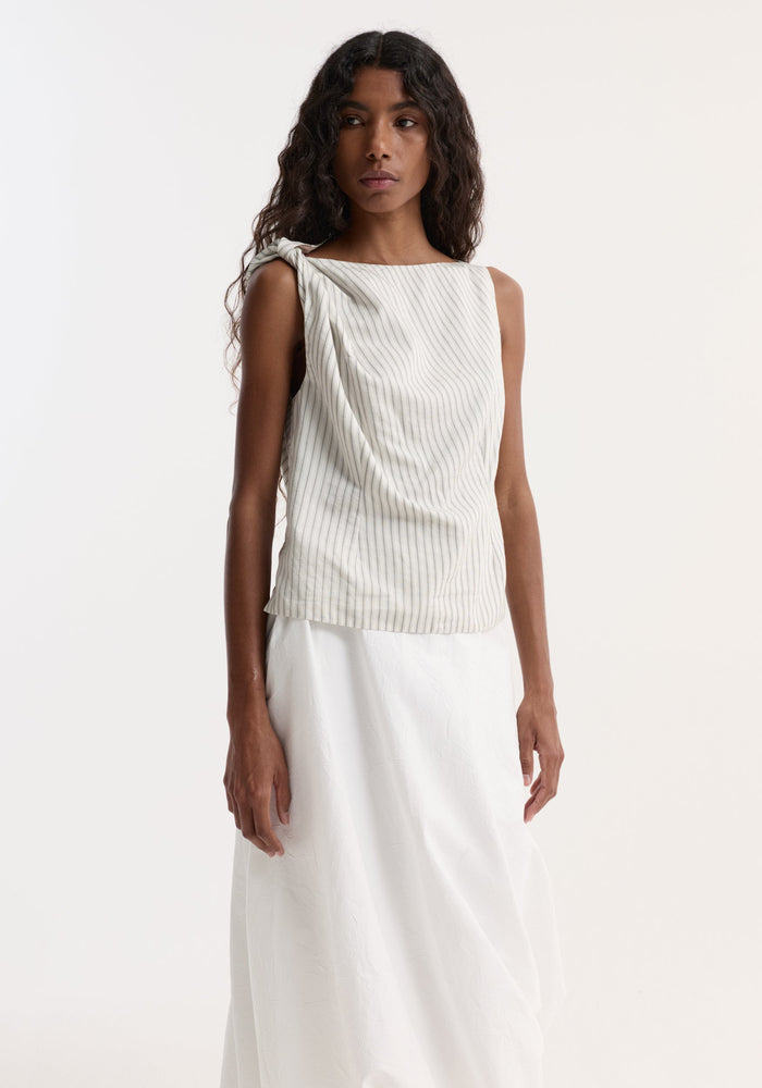 Model wearing Silk-blend draped top | fine stripe with twist detail at shoulder and boat neck, paired with a long white skirt, showcasing an asymmetrical silhouette.