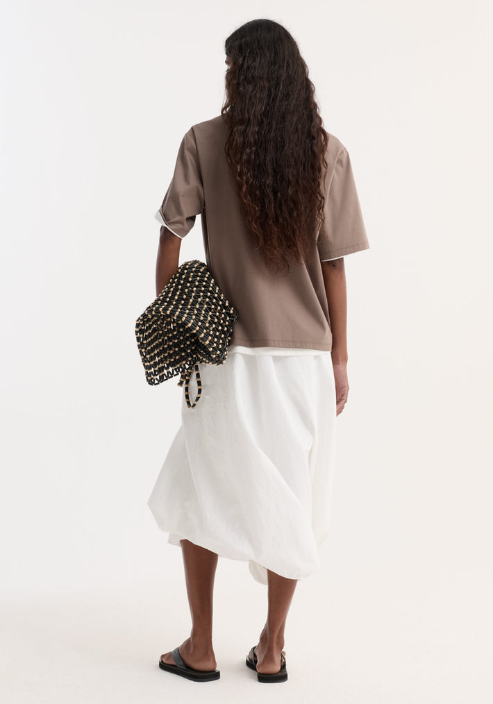 Back view of a person wearing an oversized taupe cotton-silk t-shirt with rolled sleeves, paired with a white midi skirt and black sandals.