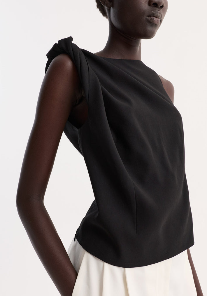 Draped linen silk top in black with a twist detail on one shoulder, boat neck, and gathered waist, creating an asymmetrical silhouette on a dark-skinned model.