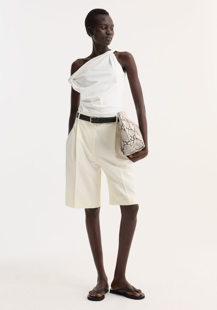Dark-skinned model wearing a Sculptural cotton silk top | off white with a one-shoulder twist detail, paired with cream shorts, black belt, sandals, and snakeskin clutch.