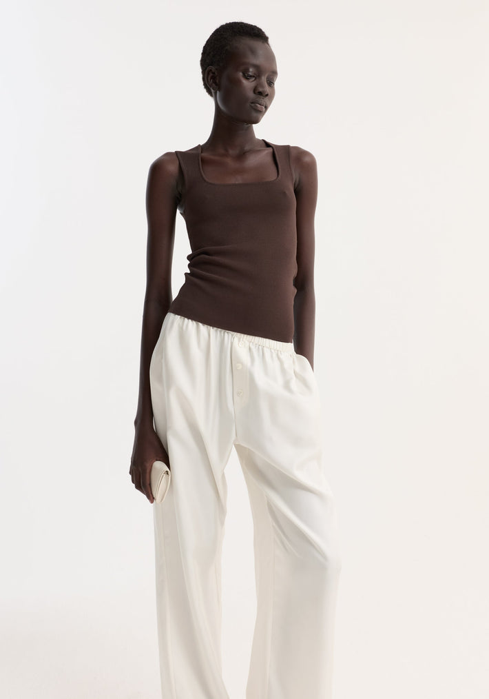 Bustier shaped knitted top in dark chocolate, featuring a fitted silhouette and sleeveless design, paired with high-waisted white trousers. The model holds a small clutch.