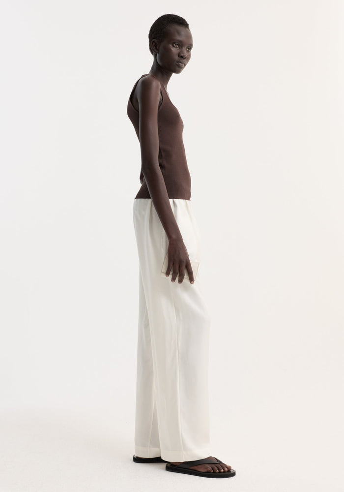 Tall person wearing Bustier shaped knitted top in dark chocolate, paired with high-waisted white trousers and black sandals, holding a neutral clutch.