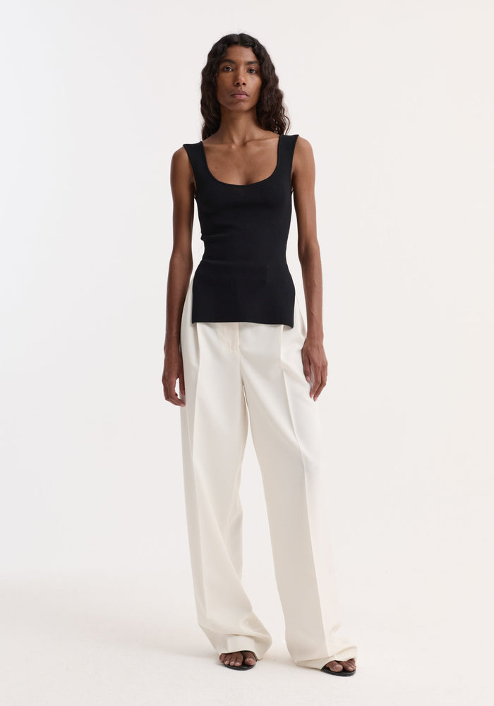 Woman wearing Long knitted bustier top with scoop neck, fitted design, and side slits, paired with high-waisted trousers and open-toe sandals, facing the camera.