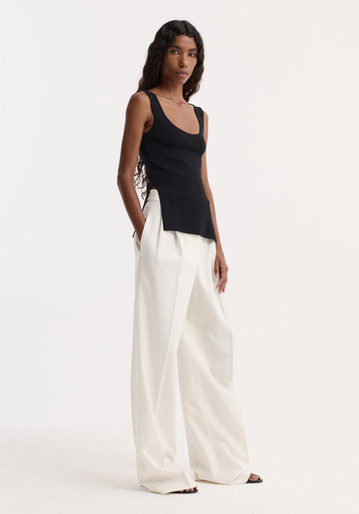 Long knitted bustier top in black with a scoop neckline, fitted design, and side splits, worn by model with high-waisted white wide-leg trousers.