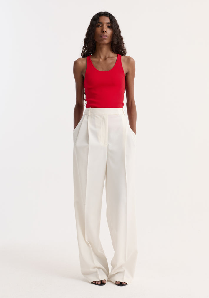 Slim person wearing a red scoop neck knitted top with high-waisted ivory trousers and black sandals, showcasing the top's fitted shape and bustier neckline.