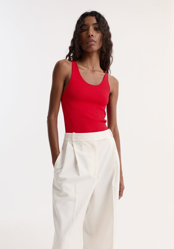 The model wears a Scoop neck knitted top | red, form-fitting with a bustier neckline, tucked into high-waisted white trousers, hands in pockets.