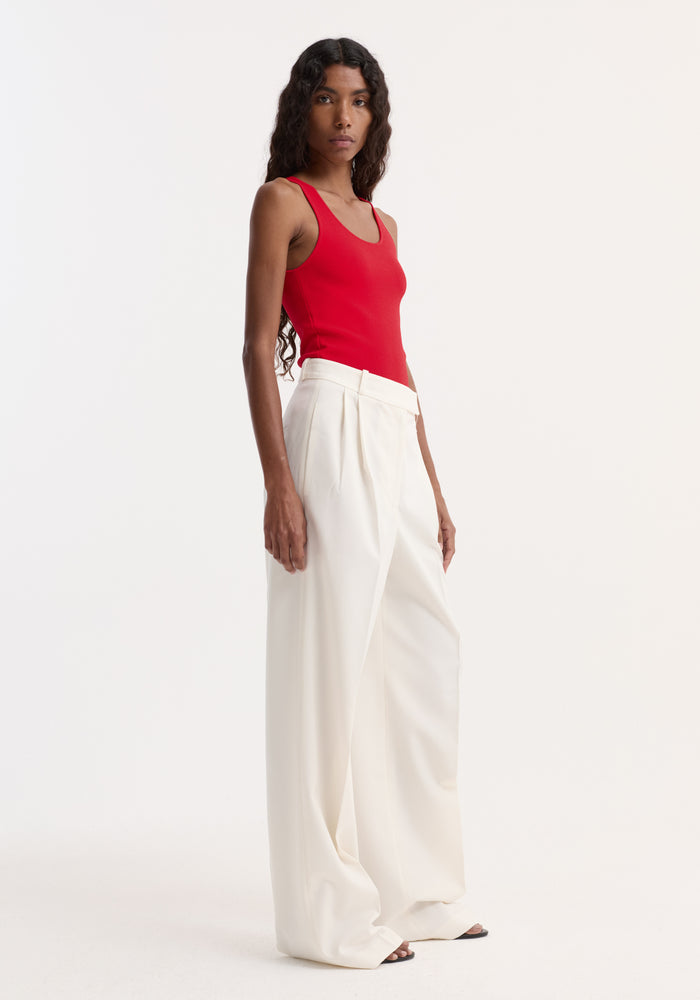 Model in a fitted, sleeveless scoop neck knitted top, red, tucked into high-waisted white trousers, showcasing the top’s bustier neckline and casual elegance.