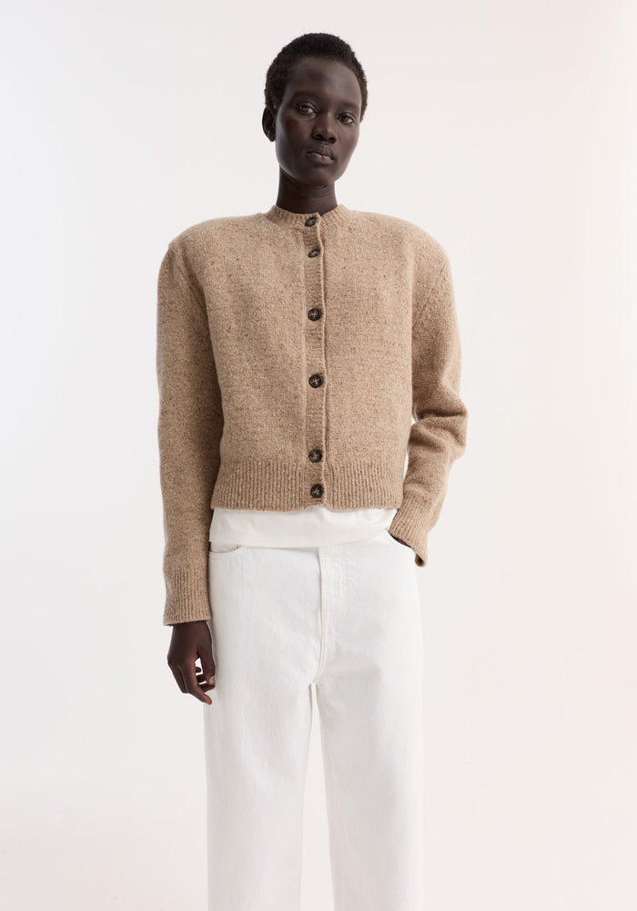 Model wearing Fine merino tweed cardigan in oat melange, featuring ribbed cuffs, dark buttons, shoulder pads, slightly cropped fit, over white top and trousers.