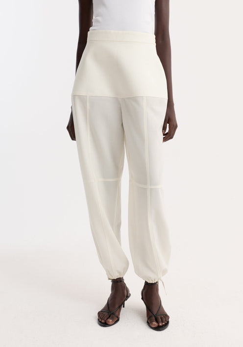 Model wearing Sculptural trousers in off white, featuring a fitted waist, seam detailing, and adjustable hems, paired with a white tank top and black sandals.