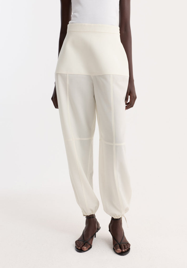 Sculptural trousers in off-white with seam detailing, worn by a model. Features elastic draw-cuff ankles, panelled waistband, and black strappy sandals.