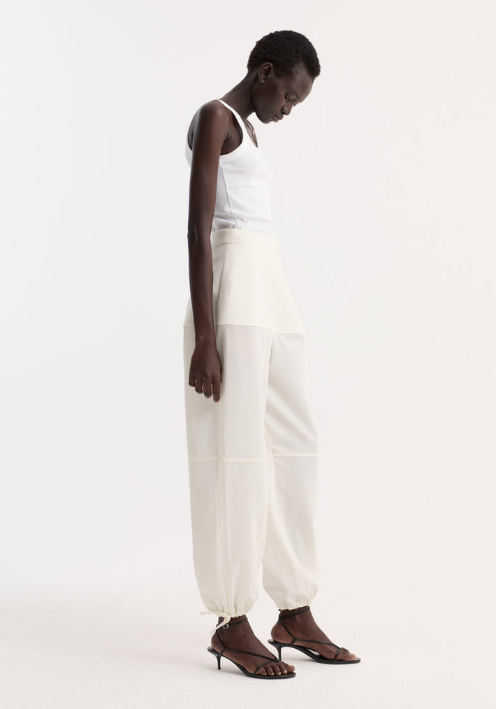 Model in profile wearing Sculptural trousers | off white, showcasing seam detailing and ankle adjusters, paired with a white ribbed tank top and black sandals.