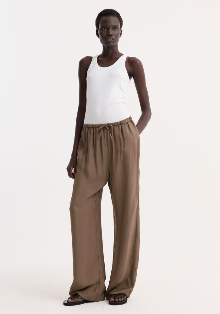 Relaxed fit trousers in brown mini check with drawstring waistband, wide legs, and side seam pockets, worn with a white ribbed tank top and sandals.