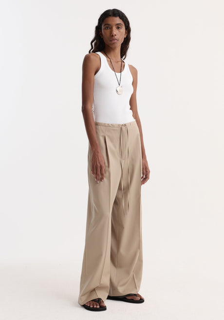 A person wears Relaxed drawstring trousers | beige, featuring a wide-leg design and drawstring waist, paired with a white tank top and black sandals.