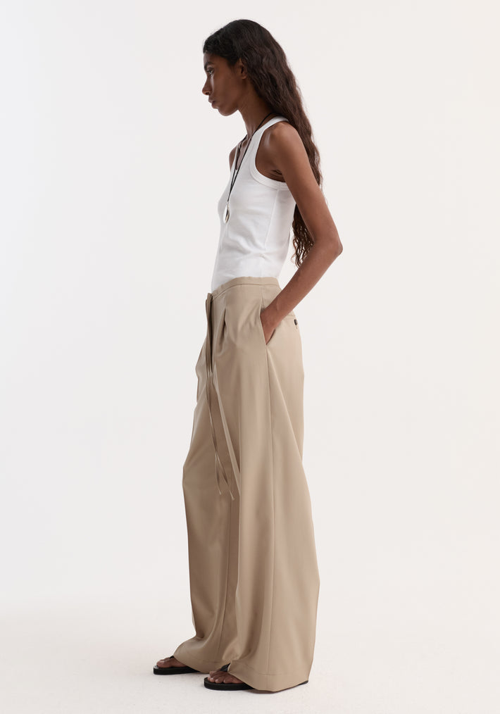 A model wears relaxed drawstring trousers in beige with a wide leg and pleats, paired with a white tank top and black sandals.