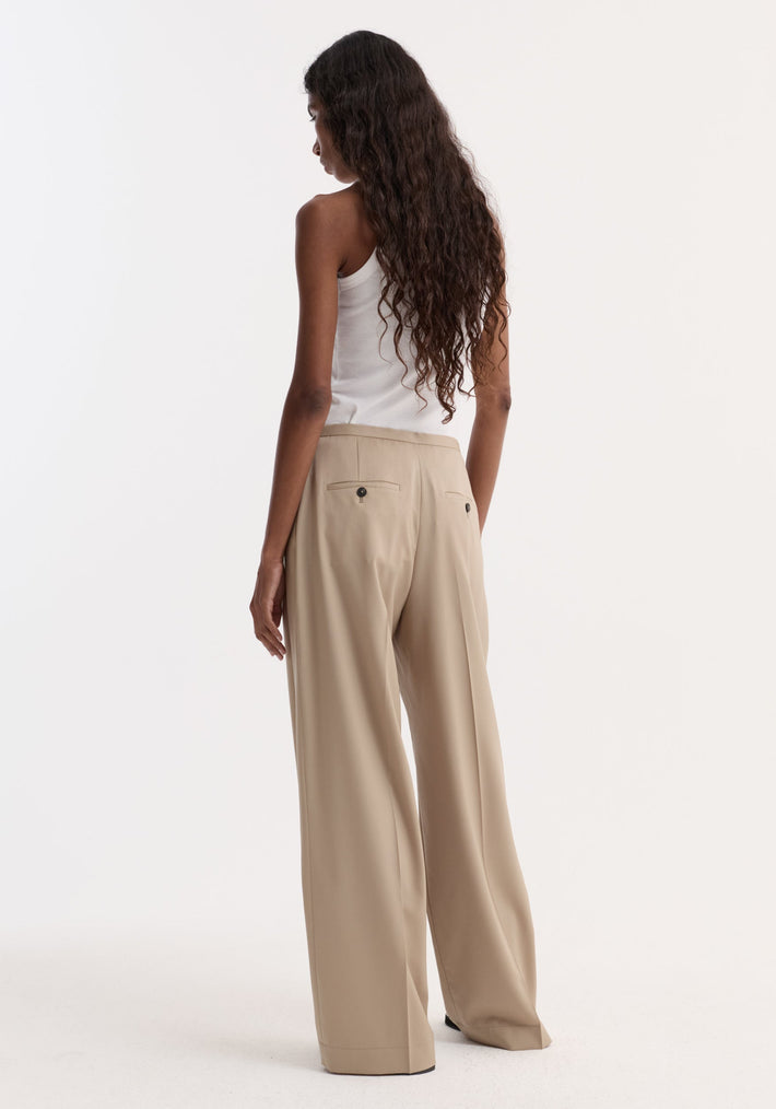 A woman in Relaxed drawstring trousers in beige, showcasing their wide-leg design and welt back pockets, paired with a sleeveless white ribbed tank top.