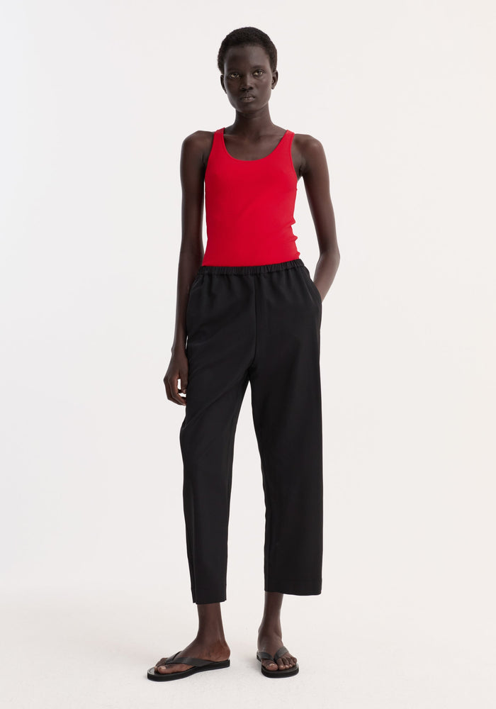 Model wearing cropped wide leg trousers in black, featuring an ankle-length fit, elasticated waist, and side pockets, paired with a red tank top.
