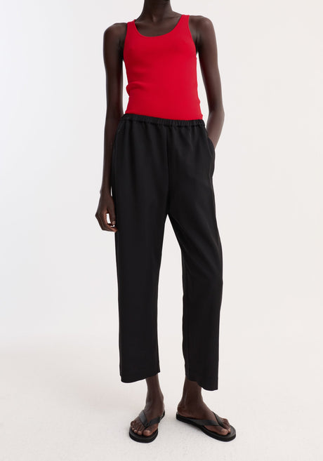 Model wearing cropped wide leg trousers in black, featuring an ankle-length fit, elasticated waist, and side pockets, paired with a red tank top.