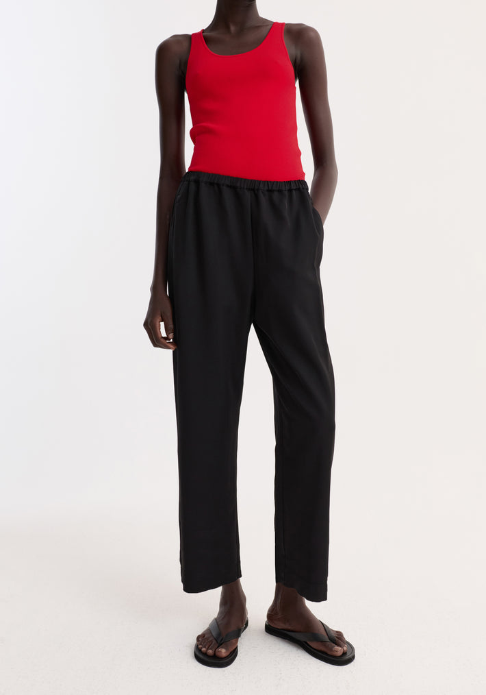 Model wearing cropped wide leg trousers in black with an elasticated waist, side seam pockets, and ankle-length fit, paired with black flip-flop sandals.