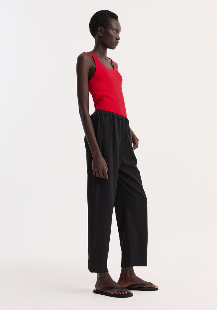Model in profile wearing black cropped wide-leg trousers with an elasticated waist and side pockets, paired with a red scoop-neck tank top and black flip-flops.