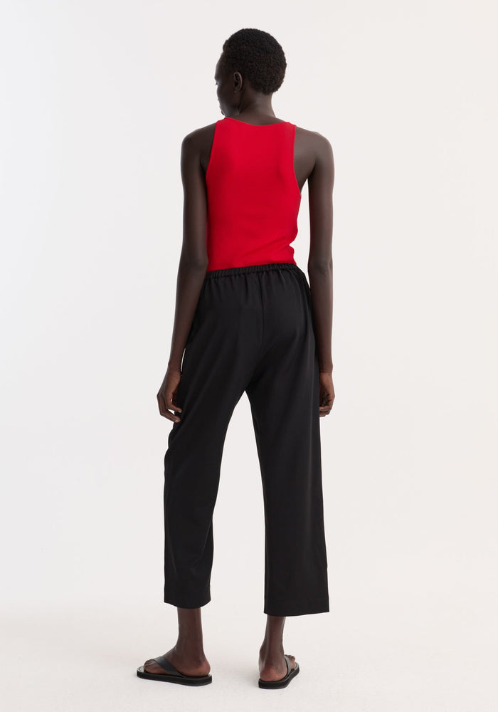 Cropped wide leg trousers in black, featuring an elasticated waist and ankle-length fit. Worn with black slide sandals, highlighting the trousers' casual elegance.