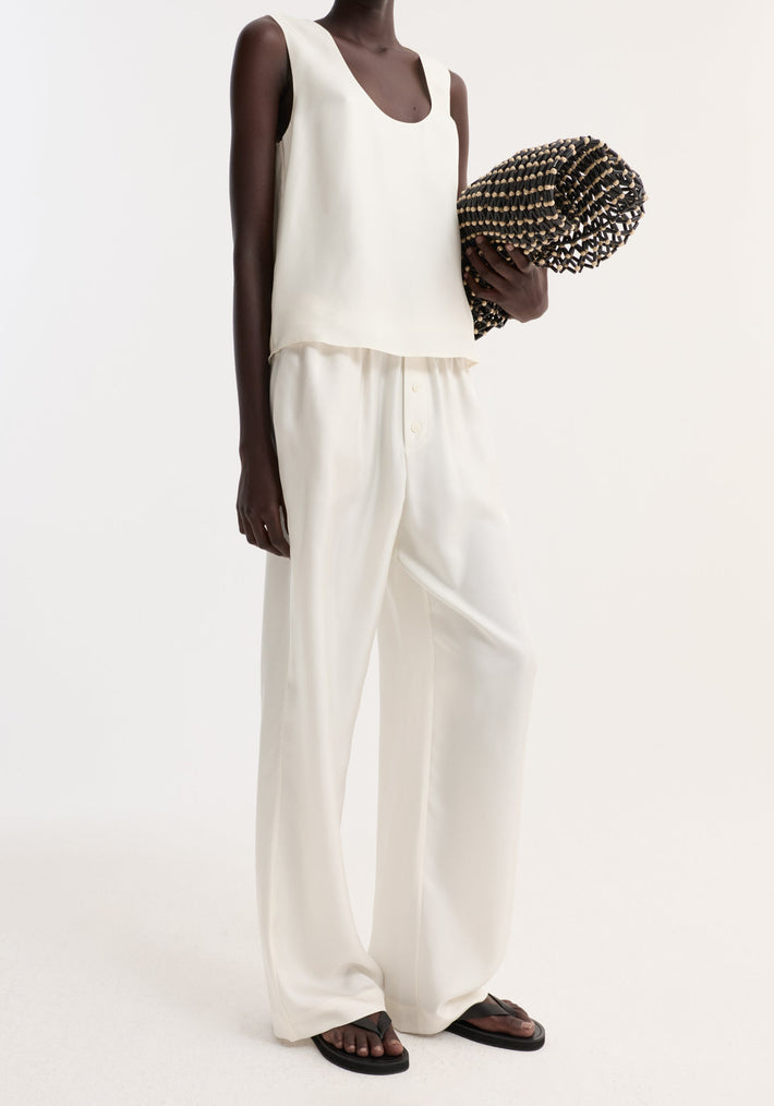 Model wearing cream Silk pyjama trousers, styled with a sleeveless silk top, holding a woven mat, and accessorized with black thong sandals.