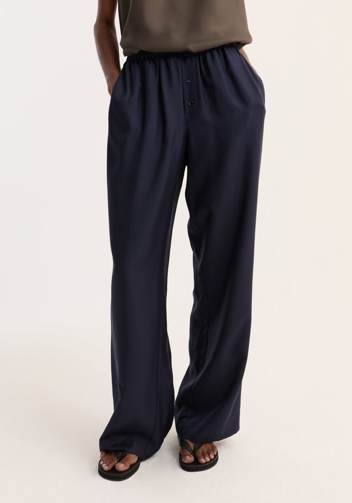 Silk pyjama trousers in navy blue, featuring wide legs, elastic waistband, and faux fly with buttons. Model wears them with sandals, hands in pockets.
