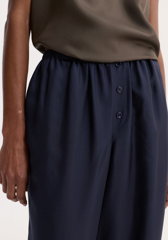 Silk pyjama trousers with elastic waistband, faux fly, two-button detail. Model's hand visible, showcasing loose-fit and luxurious silk fabric.