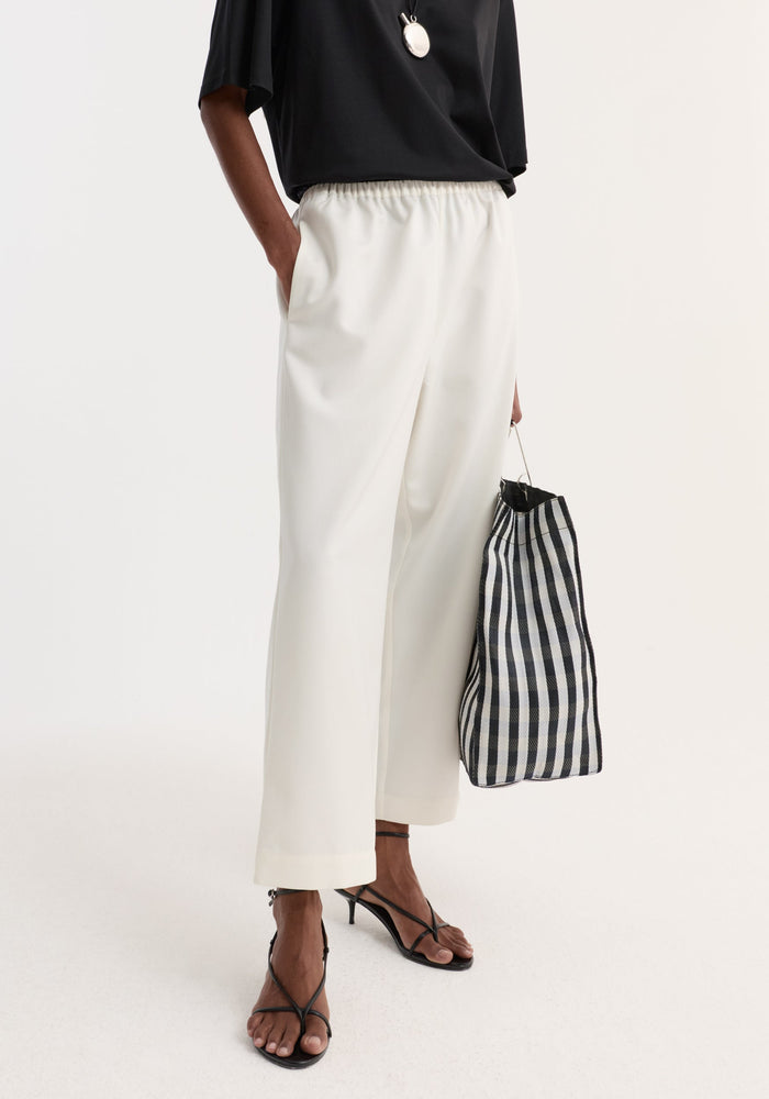 Person wearing off-white cropped wide-leg trousers with an elastic waist and side pockets, styled with black sandals and a gingham tote bag.
