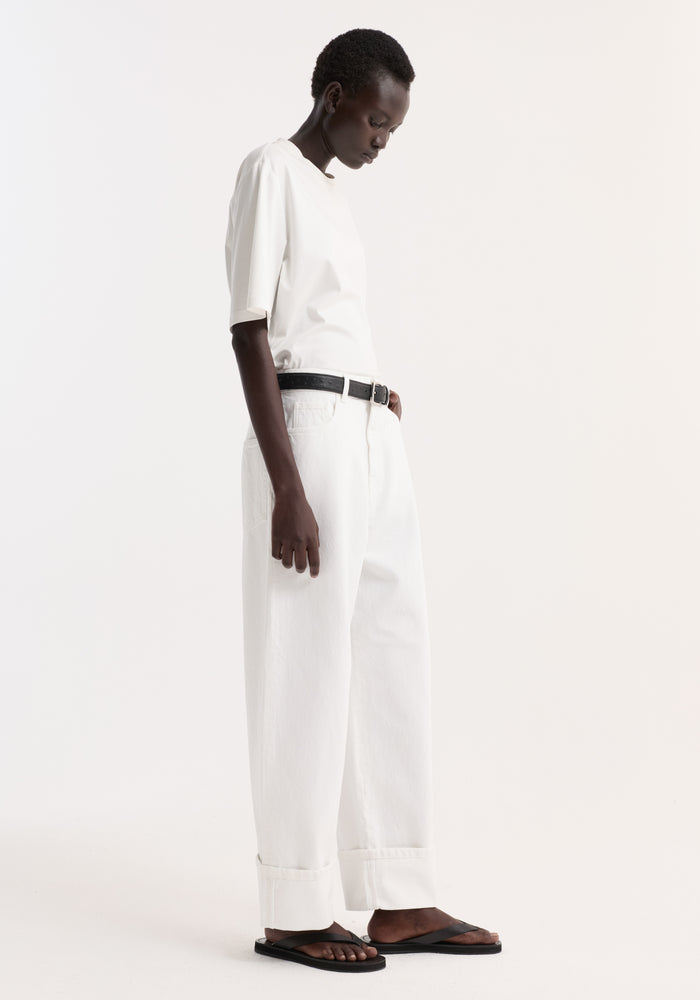 Woman in profile wearing Baggy fit turn-up denim in optic white, paired with a white T-shirt, black belt, and black thong sandals.