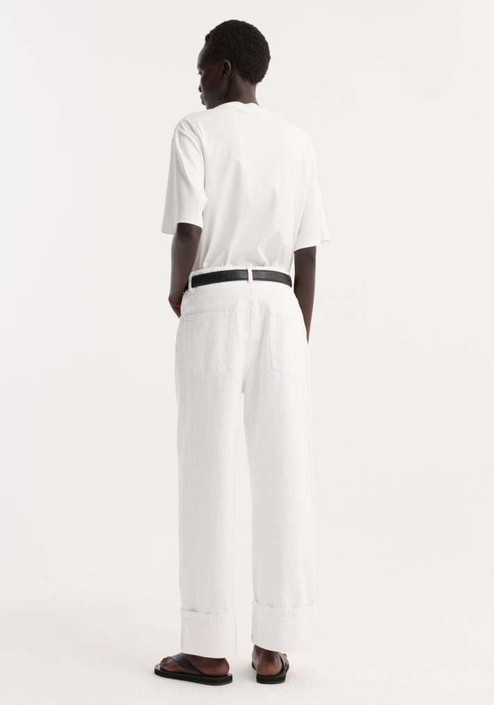 Person in Baggy fit turn-up denim | optic white, with black belt, wearing loose-fit shirt and flat sandals, showcasing minimalist style against a plain backdrop.