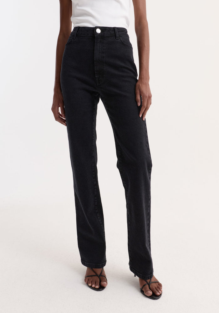 Person wearing High-rise straight stretch denim | black, featuring a slim leg and higher waist, paired with a white ribbed tank top and strappy heeled sandals.
