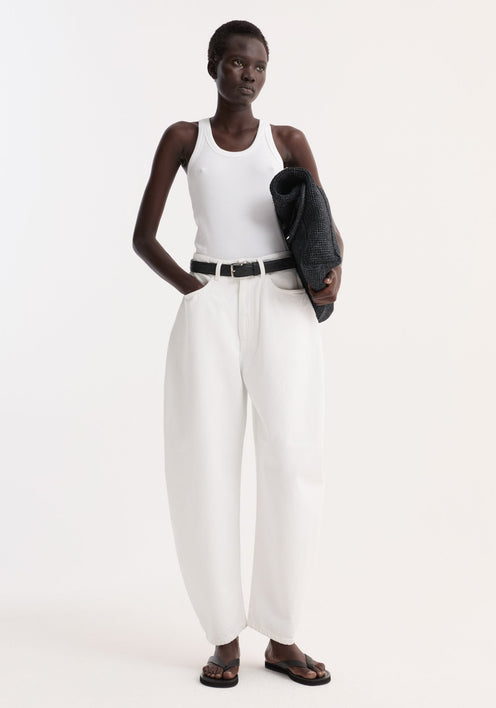 Model in optic white high-waist sculptural denim with balloon leg, paired with a white tank top and black sandals, holding a dark knit sweater.