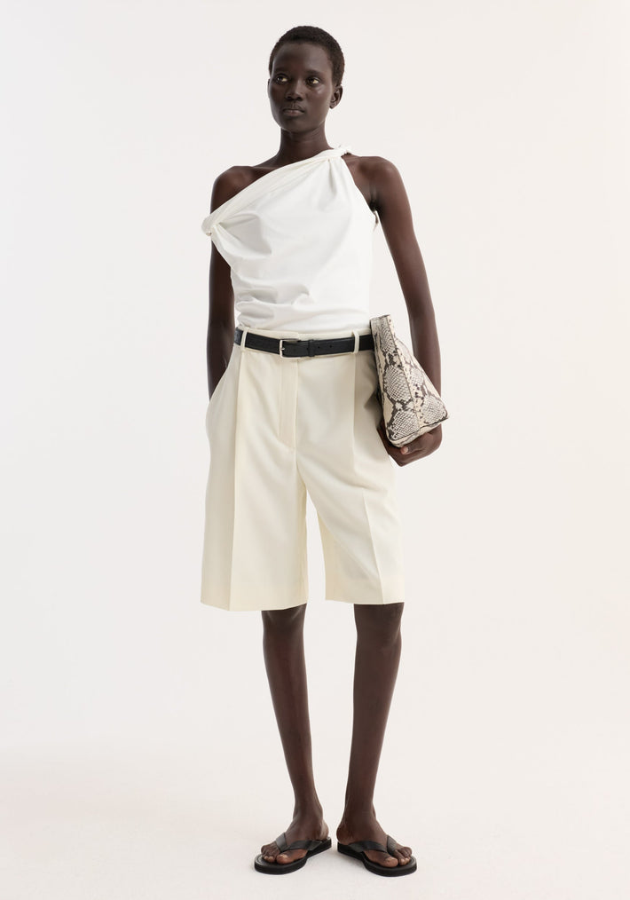 Model in Tailored single pleat shorts | off white, high-waisted with side pockets, paired with a one-shoulder top and black accessories, holding a snakeskin clutch.